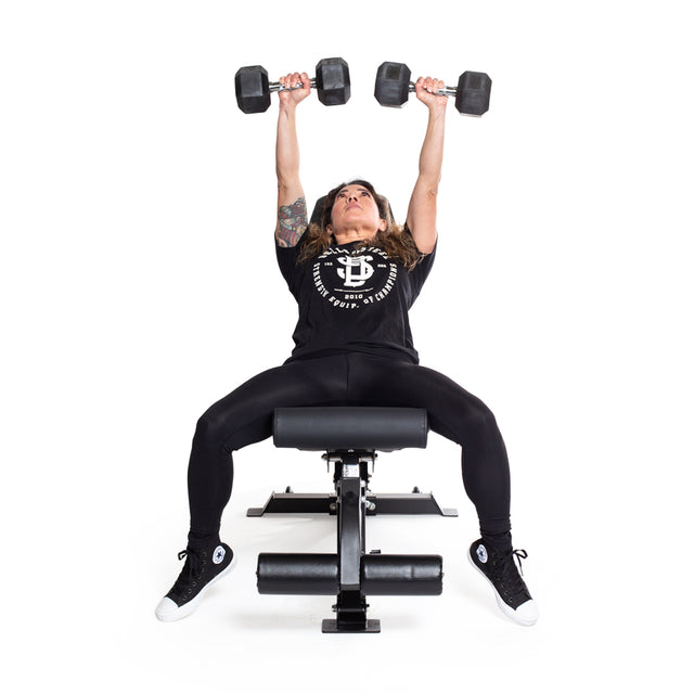 Dressed in black athletic wear, a person lifts two Bells of Steel's Ergo Rubber Hex Dumbbells overhead while lying on an exercise bench. Tattooed arms are fully extended in concentration against a plain white backdrop.