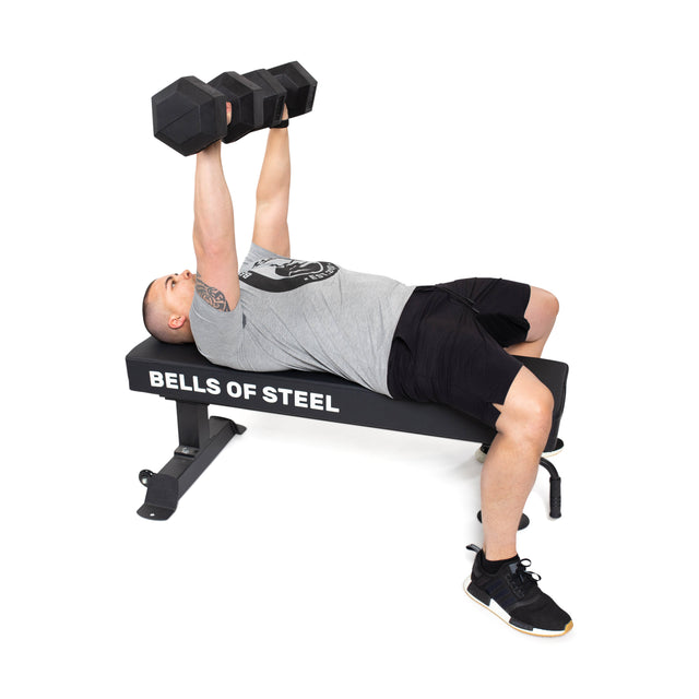 A person wearing a gray shirt, black shorts, and black sneakers with white soles is lying on a weight bench from Bells of Steel. They are doing a bench press with Ergo Rubber Hex Dumbbells that have chrome-plated handles.