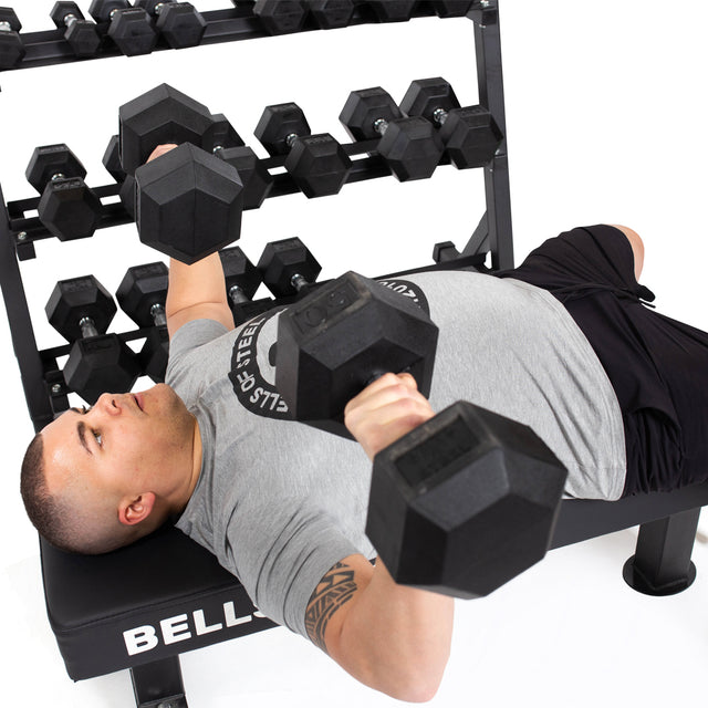 A person in a gray t-shirt and black shorts performs a chest press with two Bells of Steel Ergo Rubber Hex Dumbbells, their chrome-plated handles gleaming under the gym lights. Behind them, a rack holds more dumbbells as they focus on completing their bulk sets.