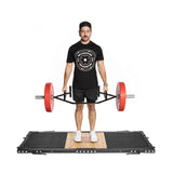 A man on a Bells of Steel Weightlifting Platform grips an Olympic barbell with red weights. He's wearing a black cap, black t-shirt with a circular logo, black shorts, and white shoes, gazing downward with a firm stance.