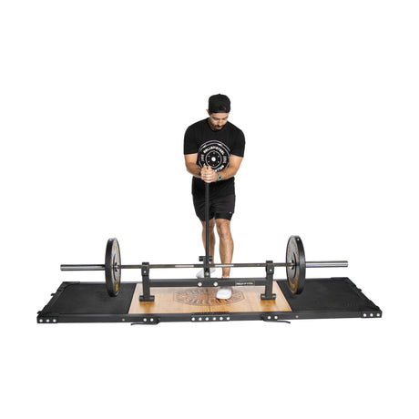 A man confidently stands on the Bells of Steel Weightlifting Platform, equipped with deadlift pads, wearing a black shirt, shorts, and cap. He expertly uses an Olympic barbell with weights on each side. The platform supports various weightlifting exercises.