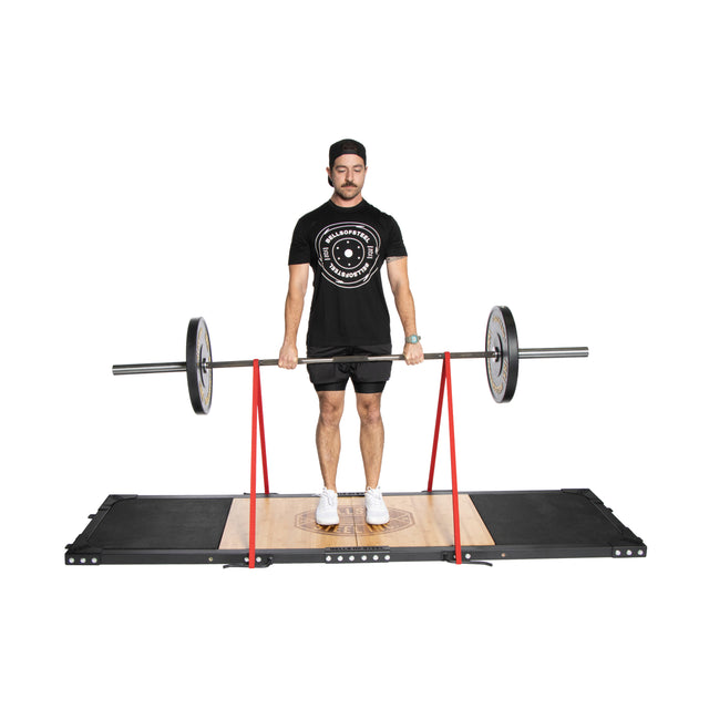 Wearing a black shirt, shorts, and cap, a man deadlifts with an Olympic barbell equipped with red support straps on the Bells of Steel Weightlifting Platform.