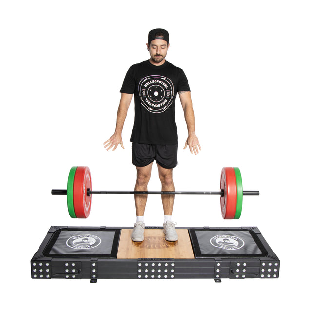 A man stands on the Bells of Steel Weightlifting Platform, eyes closed and arms down, preparing for a deadlift. He faces forward in a black shirt, shorts, and cap with white sneakers, ready before the Olympic barbell with red and green weights on sturdy deadlift pads.