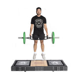 A man in athletic wear jumps mid-air while performing a deadlift with a trap bar and green plates on a Bells of Steel Weightlifting Platform. He wears a cap, black shirt, shorts, and gray sneakers. The platform is marked with a logo and geometric design.