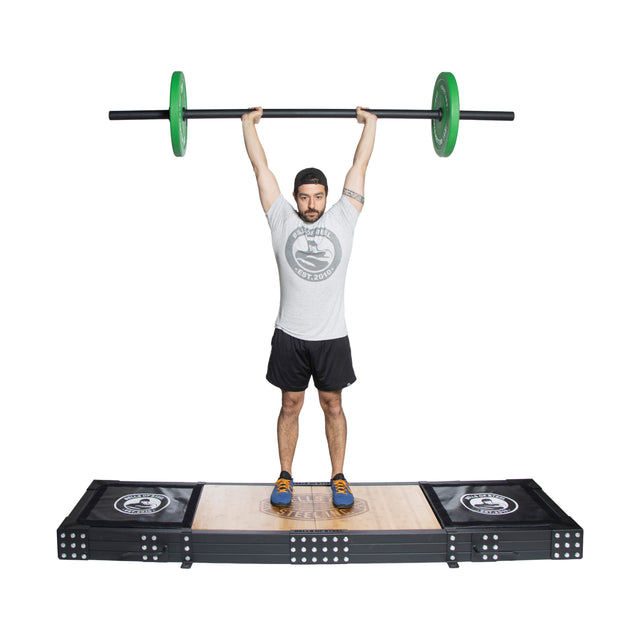 Wearing a gray shirt, black shorts, and yellow shoes, an individual stands on the Bells of Steel Weightlifting Platform, lifting an Olympic barbell with green weights overhead with a focused expression.