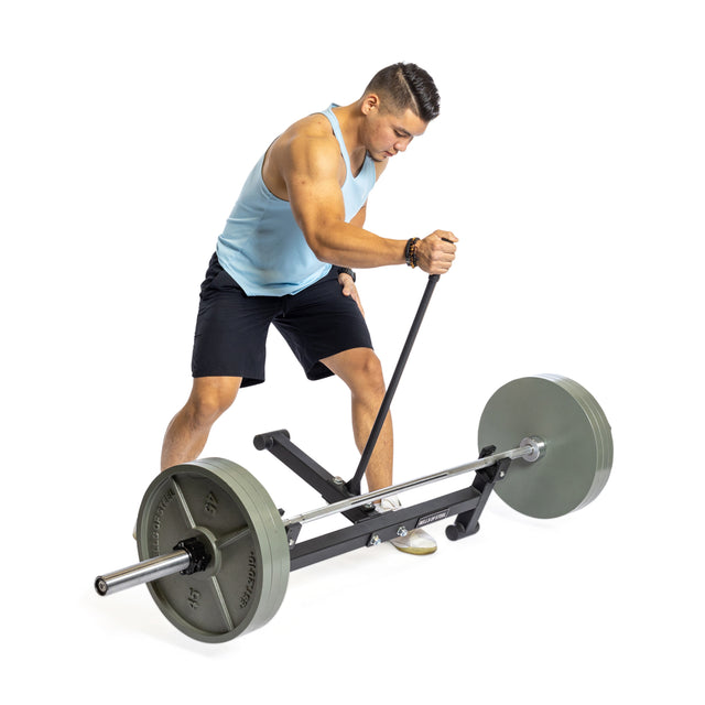 A person wearing a light blue tank top and black shorts performs a bent-over row using a Bells of Steel Deadlift Jack with Rollers, set up for weight changes. The individual is on a white background, concentrating on the exercise.