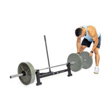 A person in a light blue tank top and black shorts uses the Bells of Steel Deadlift Jack with Rollers to load weights onto a barbell on a gym rack, carefully adding a weight plate to one side for smooth, precise changes.