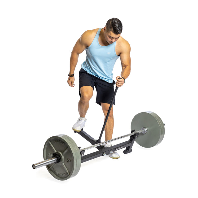 A person wearing a blue tank top and black shorts prepares to lift a loaded barbell using the Bells of Steel Deadlift Jack with Rollers. They are surrounded by nylon rollers in a brightly lit setting on a white background.