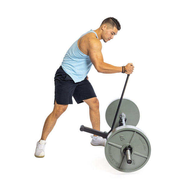 A man in a light blue tank top and black shorts exercises using a barbell with weight plates on a landmine attachment, showcasing the versatility of the Bells of Steel Deadlift Jack with Rollers. He leans forward, gripping the bar against a plain white background.