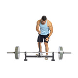 Wearing a blue tank top and black shorts, a person adjusts a heavy barbell on a squat rack. They use the Bells of Steel Deadlift Jack with Rollers to streamline weight changes, focusing intently. The plain white background emphasizes their dedication.