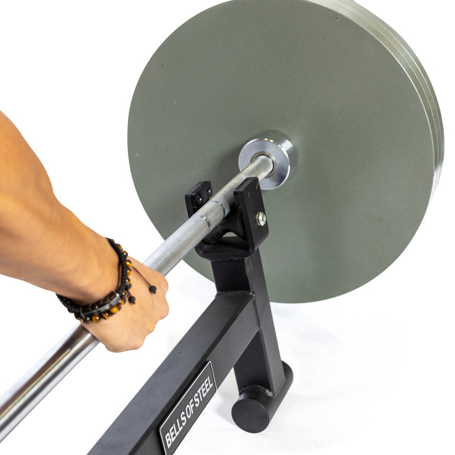 A person grips the handle of the trap hex bar, loaded with heavy circular weights on a black metal frame labeled "Deadlift Jack with Rollers" by Bells of Steel, featuring smooth nylon rollers for effortless maneuverability and boasting an impressive 800lb capacity.