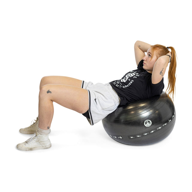 A person with long red hair performs crunches on a Bells of Steel Exercise Ball. Wearing a black shirt, gray shorts, white socks, and white sneakers against a plain white background, they display perfect form and focus in their workout routine.