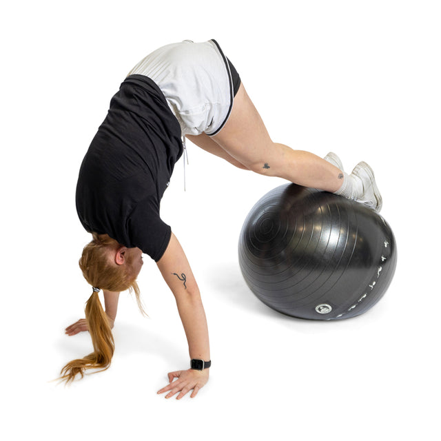 A person with long hair in a ponytail performs a pike exercise using a large black Bells of Steel Exercise Ball for support. They are dressed in a black shirt, light shorts, and white sneakers against a plain white backdrop.