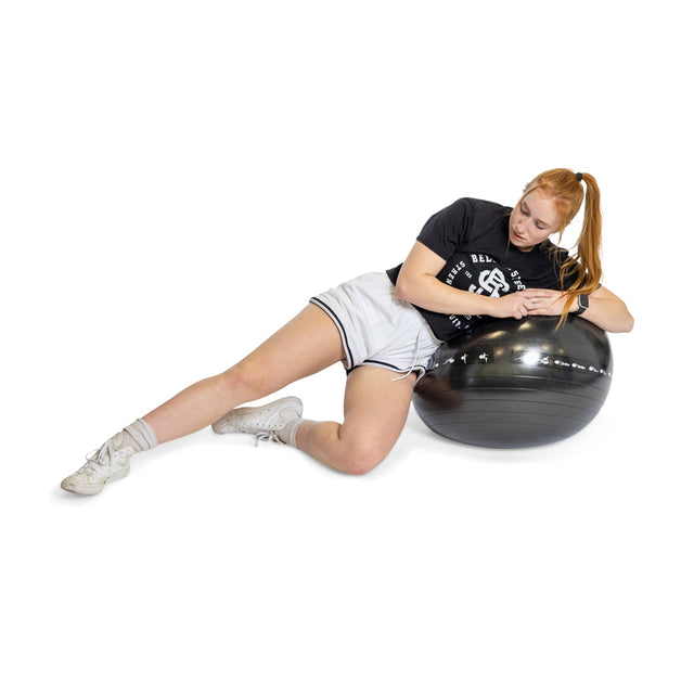 A red-haired person integrates the Bells of Steel Exercise Ball into their workout, lying sideways on the black ball. They wear a black shirt, white shorts, socks, and white sneakers with hands resting on the ball against a plain white background.