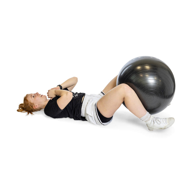 With red hair and dressed in a black t-shirt, white shorts, and sneakers, a person lies on their back with knees bent, holding a Bells of Steel Exercise Ball between their legs during a workout against a plain white background.