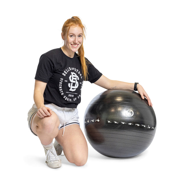 A person with long red hair smiles while kneeling on the floor, dressed in a black T-shirt, gray shorts, and white sneakers. Their left hand rests on a Bells of Steel Exercise Ball, showcasing ideal workout equipment for balance training.