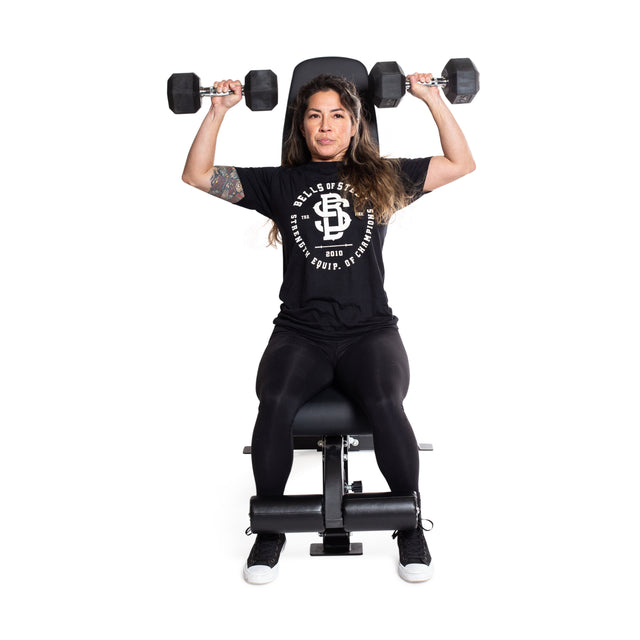 A person is seated on the Bells of Steel Flat / Incline / Decline Weight Bench - Commercial, performing a shoulder press with two dumbbells. They are wearing a black T-shirt with a white logo, black leggings, and black sneakers against a plain white background.