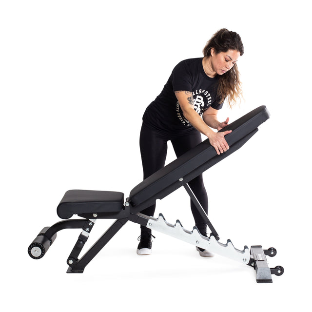 A woman wearing a black t-shirt, black leggings, and sneakers is adjusting the backrest of a versatile Bells of Steel Flat / Incline / Decline Weight Bench - Commercial. The bench has a padded seat and backrest, supported by a heavy-duty metal frame with adjustable incline settings.