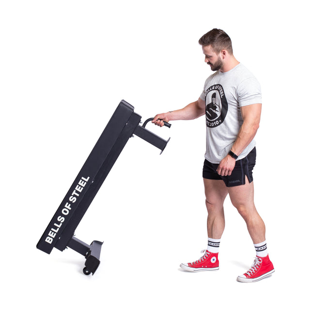 A man in a T-shirt and shorts tilts the Bells of Steel Powerlifting Flat Bench on wheels, wearing red sneakers and socks. Set against a white background, this exercise equipment boasts an impressive 1,000lb max capacity.