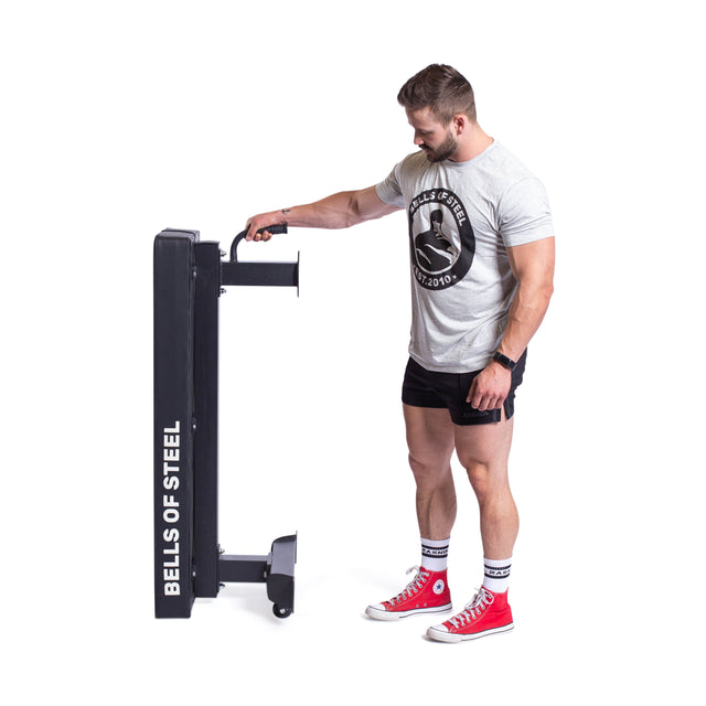 A man in a gray "Bells of Steel" shirt and red sneakers sets up a branded "BELLS OF STEEL" Powerlifting Flat Bench on a white background, confidently preparing for exercise with its impressive 1,000lb max capacity.