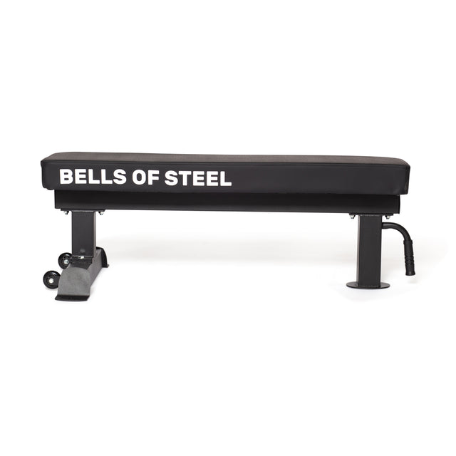 This light commercial grade Powerlifting Flat Bench by Bells of Steel features a black padded top, two supporting black legs, wheels for easy transport, and a 1,000lb max capacity, all set on plain white flooring.