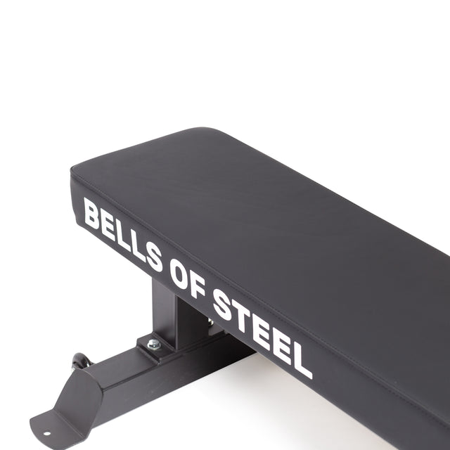 A close-up shows the Bells of Steel Powerlifting Flat Bench, with "Bells of Steel" in white on its side. This bench is engineered for light commercial use and stands robustly on a sturdy black metal frame.