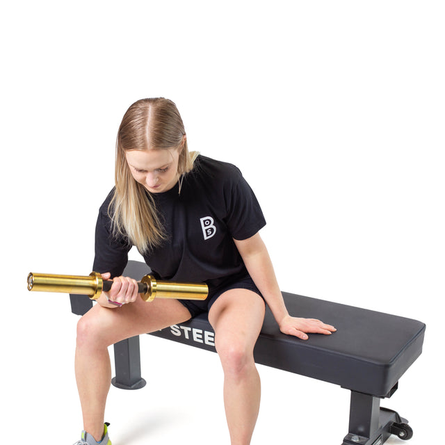 A person in a black shirt, shorts, and athletic shoes sits on the Bells of Steel Powerlifting Flat Bench, doing a one-handed barbell curl. The sturdy bench supports up to 1,000 lb against a plain white backdrop as they showcase impressive strength.