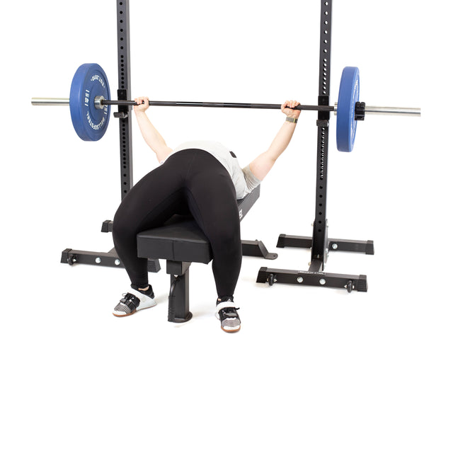 A person uses the Bells of Steel Powerlifting Flat Bench to bench press with a barbell loaded with blue plates, suited for light commercial use. They are in black leggings, a light shirt, and athletic shoes against a white background. The setup supports up to 000lb max capacity.