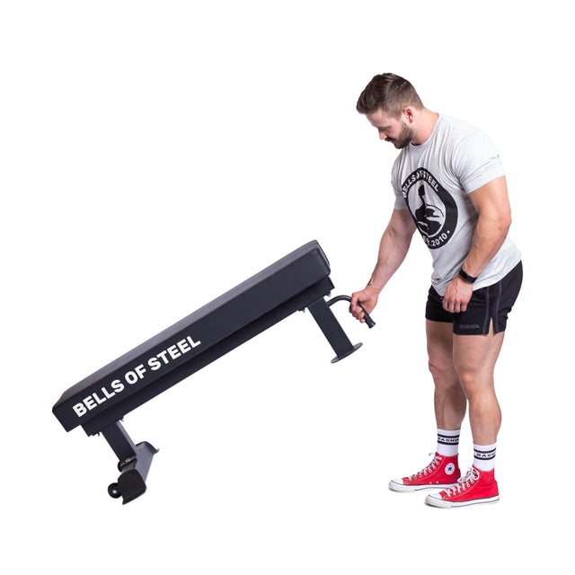 A person in red sneakers and gym attire adjusts a Bells of Steel Powerlifting Flat Bench, recognized for its light commercial grade quality. The bench is set upright, with the individual positioned against a white background.