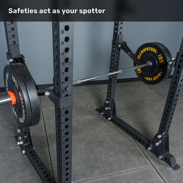 A Bells of Steel Manticore Flat Foot Power Rack Builder (3" x 3", 1" Holes) with weight plates rests on the safety bars in a squat rack. Text reads, "Safeties act as your spotter." The tiled gym floor and numbered adjustment holes make this 3x3 power rack perfect for any home gym setup.