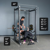 Two people are in a gym, with one bench pressing a barbell inside the Bells of Steel Manticore Flat Foot Power Rack Builder (3" x 3", 1" Holes) while the other spots. This sturdy power rack is ideal for any home gym, offering clearly marked dimensions: width 49", height options of 75¼", 87¼", 93¼", or 111¼", and depth options of 54" or 63".