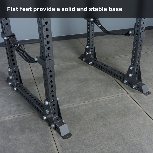 Close-up of the Bells of Steel Manticore Flat Foot Power Rack - Prebuilt (3" x 3", 1" Holes), featuring sturdy flat feet on a tiled gym floor—ideal for any home gym setup. Text at the top reads, "Flat feet provide a solid and stable base.