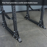 Close-up of the black metal flat feet of the Bells of Steel Manticore Flat Foot Power Rack - Prebuilt (3" x 3", 1" Holes) on a gray tiled floor, featuring holes for adjustment. Text at the top reads, “Flat feet provide a solid and stable base,” making this power rack an ideal addition to any home gym.