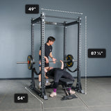 Two people use a Bells of Steel Manticore Flat Foot Power Rack - Prebuilt (3" x 3", 1" holes) for bench pressing in a home gym. The rack’s dimensions are marked: 49" wide, 54" deep, and 87¼" tall. One person is lying on a bench pressing a barbell while the other stands nearby spotting.
