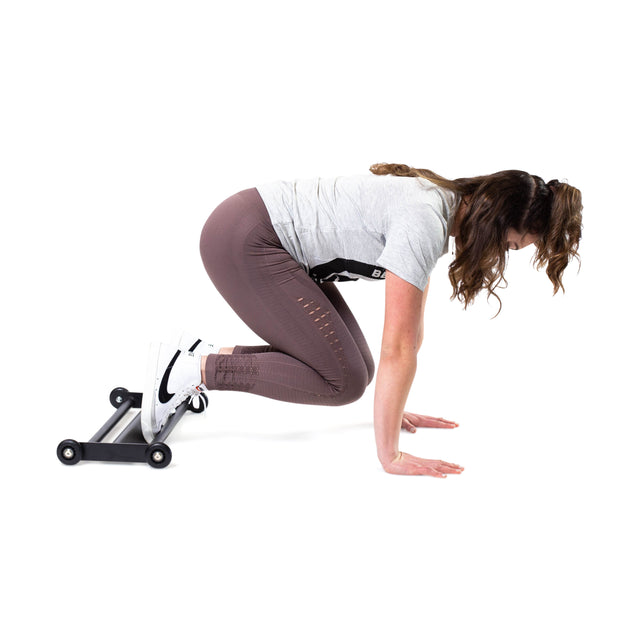 An individual in athletic wear performs a hamstring-blasting exercise on a compact fitness device, the Bells of Steel Glute Ham Slider. They balance on hands and feet with bent knees against a plain white backdrop.