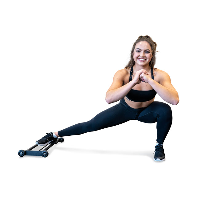 A woman in black athletic wear, featuring a sports bra, leggings, and sneakers, performs a side lunge while smiling. She uses the Bells of Steel Glute Ham Slider under her foot for an intense hamstring workout against a white background.