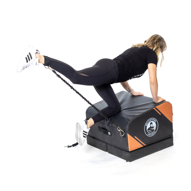 A woman in athletic wear uses the Bells of Steel Soft Glute Bench and a resistance band to perform a leg extension exercise at home, supporting herself on her hands while extending one leg back.