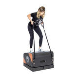 A woman in athletic wear stands on the Bells of Steel Soft Glute Bench, using resistance bands attached to its sides, exercising against a plain white background.