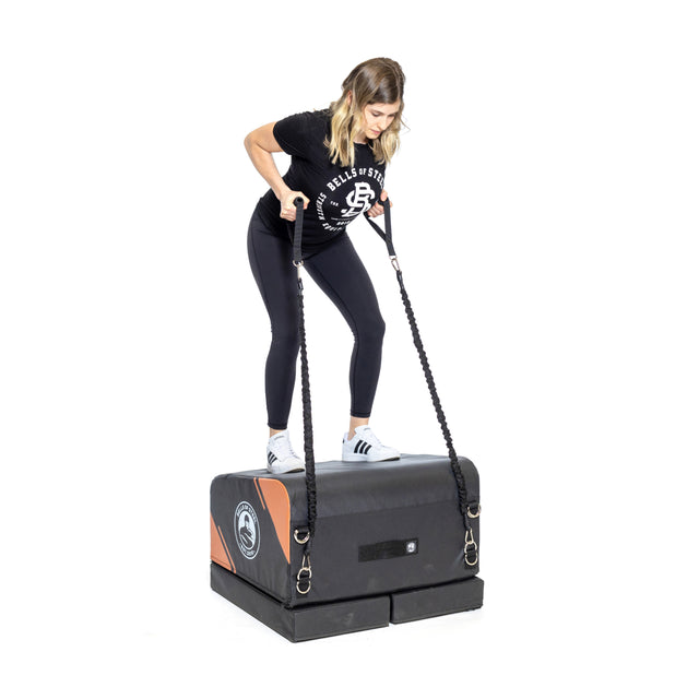 A woman in athletic wear stands on the Bells of Steel Soft Glute Bench, using resistance bands attached to its sides, exercising against a plain white background.