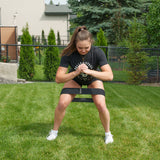 A person wearing a black shirt and shorts exercises outdoors with the Bells of Steel Glute Loop resistance band positioned above the knees, squatting on grass with hands clasped in front, in a fenced backyard for an effective booty workout.