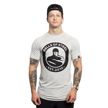 A young man in a backward black cap and a light grey Bells of Steel Bamboo T-Shirt stands against a white background, displaying tattoos on both arms and a neutral expression.