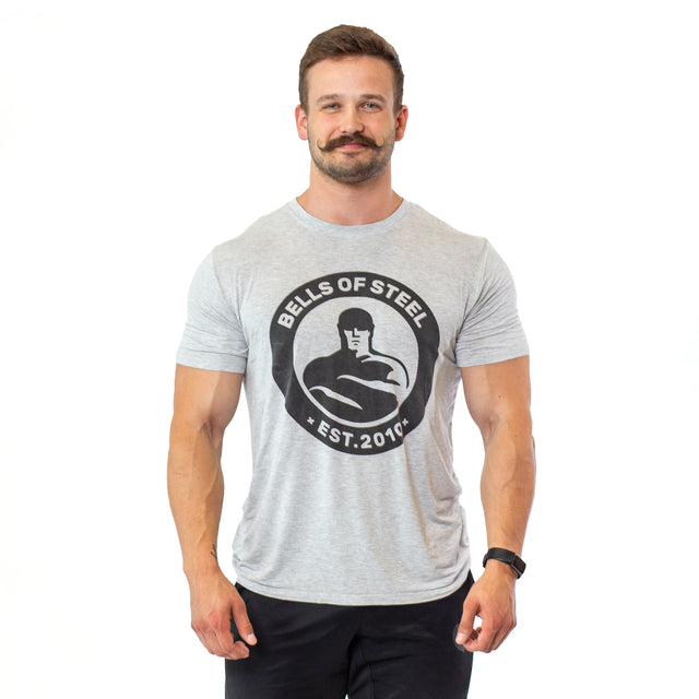 A mustachioed man stands against a white background, wearing dark pants and a watch. He showcases comfort and style in a buttery soft gray Bamboo T-Shirt from Bells of Steel, featuring the brand logo and "EST. 2010" text.