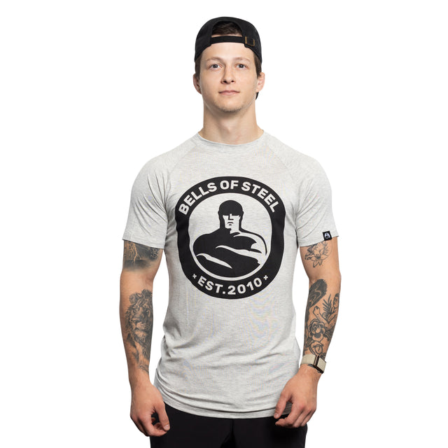 A tattooed individual stands against a white background, wearing a buttery soft "Bells of Steel" Bamboo T-Shirt with a large black logo featuring a muscular figure and "EST. 2010." They sport a backward baseball cap.