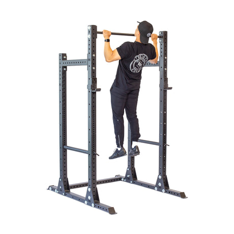 Wearing black athletic gear and a cap, an individual performs a pull-up on the Bells of Steel Hydra Half Rack—Prebuilt (3" x 3", ⅝" Holes), utilizing its modular components for enhanced versatility.