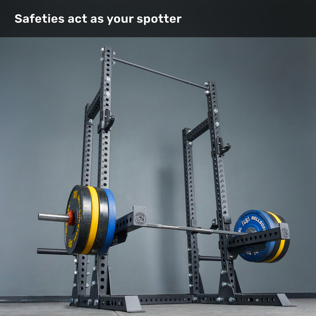 A loaded barbell rests on the Bells of Steel Manticore Half Rack Builder (3" x 3", 1" Holes) with sturdy uprights and safety bars. Text above reads, “Safeties act as your spotter.” The plain gray background makes it a perfect fit for any home gym or Half Rack setup.
