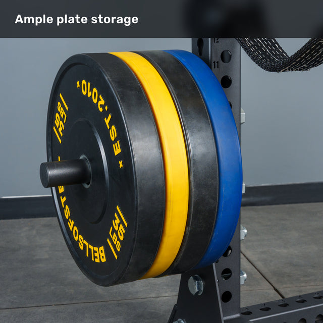 Three large weight plates—black, yellow, and blue—are stacked on a gym plate holder attached to the Bells of Steel Manticore Half Rack Builder (3" x 3", 1" Holes), whose robust uprights make it an excellent addition to any home gym. The text "Ample plate storage" appears at the top.