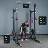 A person lifts a barbell inside the Bells of Steel Manticore Half Rack Builder, which features sturdy 3" x 3" uprights with 1" holes. White dotted lines and measurements highlight the rack’s height options (87¼” or 93¼”), width (49”), and depth choices (54” or 63”). Designed as the perfect addition to any home gym, this versatile half rack also showcases convenient gym accessories hanging from its frame.