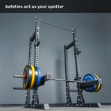 A loaded barbell rests on the safeties of the Bells of Steel Manticore Half Rack - Prebuilt (3" x 3", 1" Holes), set against a gray wall, with the caption “Safeties act as your spotter” above—making it an ideal addition to any home gym setup.