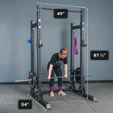 A person prepares to lift a barbell inside the Bells of Steel Manticore Half Rack – Prebuilt, which features robust 3" x 3" uprights with 1" holes. The rack’s dimensions are labeled: 87¼ inches tall, 49 inches wide, and 54 inches deep. Weight plates and resistance bands hang on the Manticore Half Rack, making it an ideal addition to any home gym.