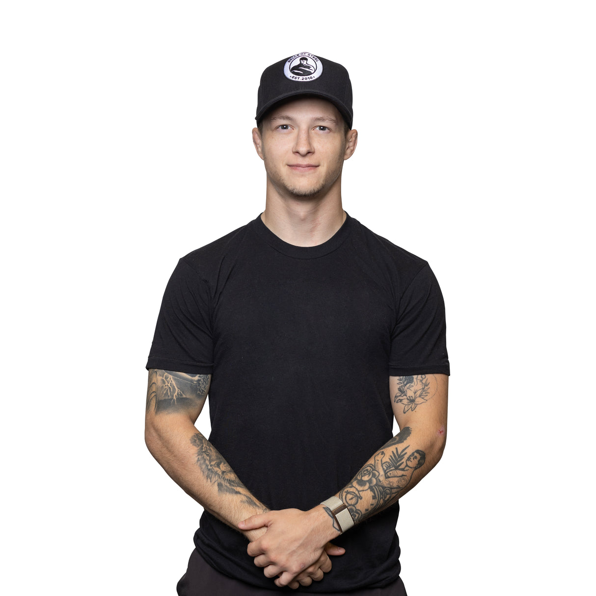 A young man wearing the Bells of Steel Snap Back Hat and a black t-shirt stands against a white background, showing off his arm tattoos and smiling at the camera with his hands clasped in front.